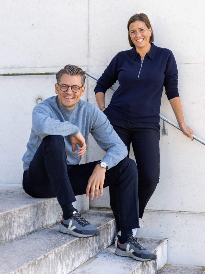 Foto von Simone Brehm (stehend) und Markus Brehm (sitzend) auf Treppe, beide lächeln in die Kamera
